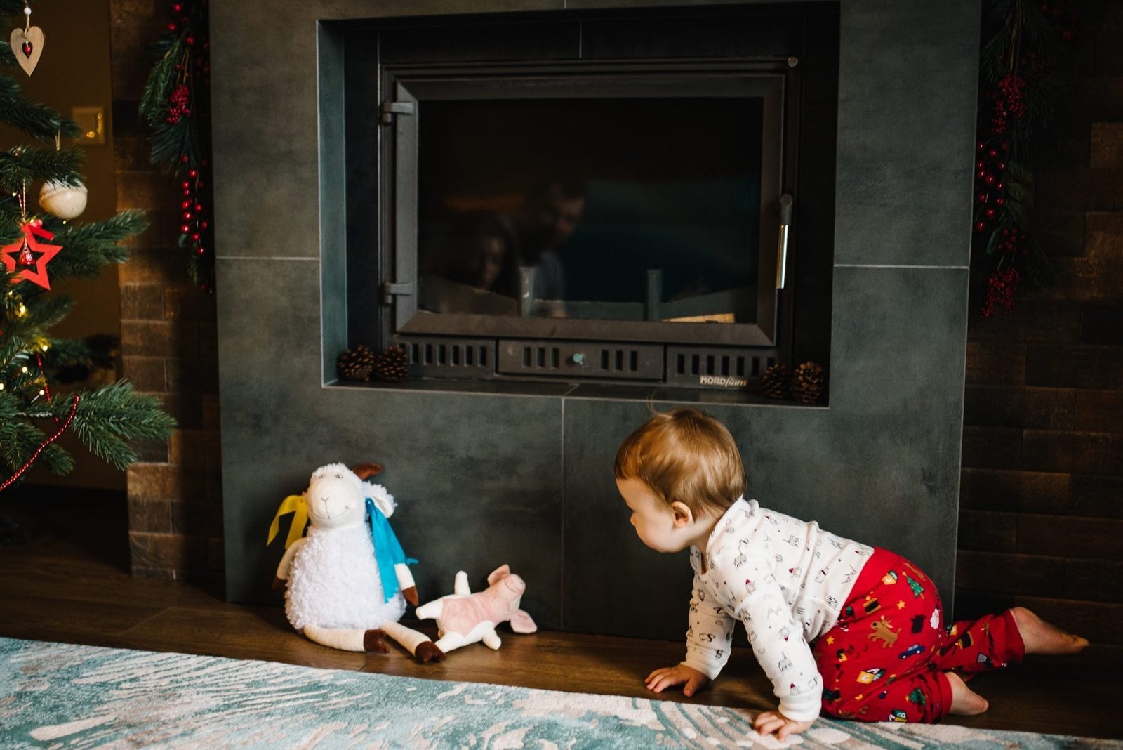 A child crawls toward two stuffed animals near a fireplace, surrounded by festive decorations and a Christmas tree, creating a cozy indoor a