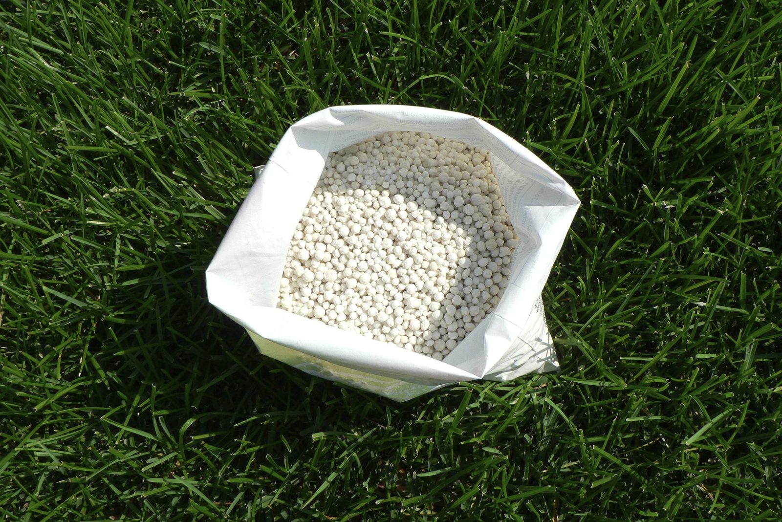 A bag filled with small white pellets rests on green grass, suggesting use in gardening or fertilization. The bag is partially open at the t