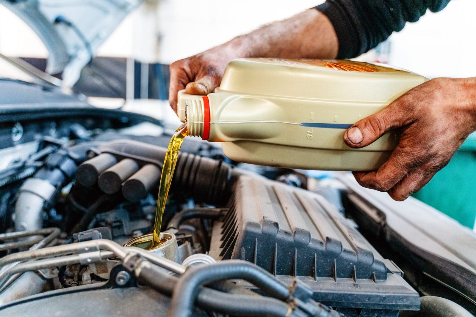 A person pours engine oil from a container into a vehicle's engine compartment, surrounded by mechanical components in a garage setting.