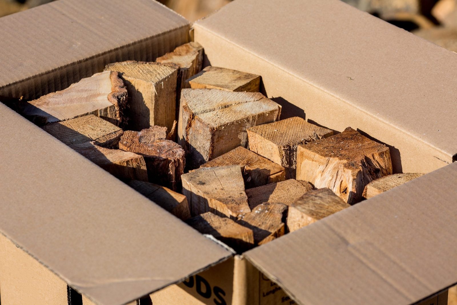 A cardboard box holds neatly arranged pieces of firewood, indicating preparation for use in a fireplace or campfire, set in a bright outdoor