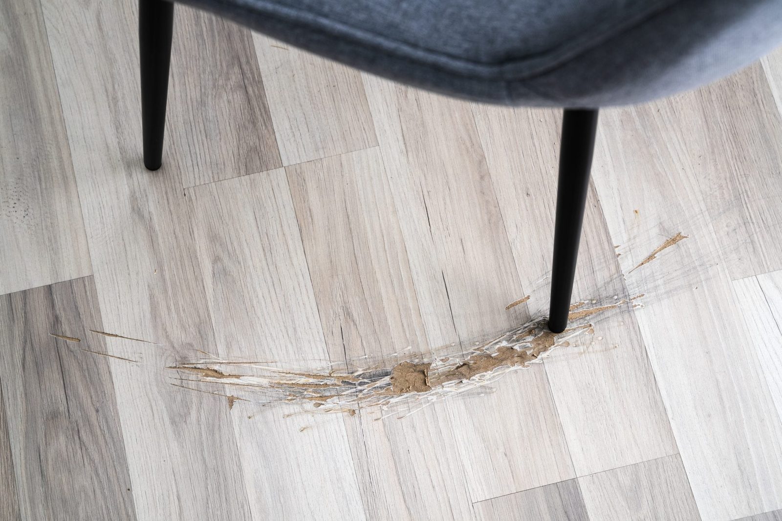 A black chair leg rests on scratched hardwood flooring, revealing distinct marks and discolored spots around the base, indicating wear from