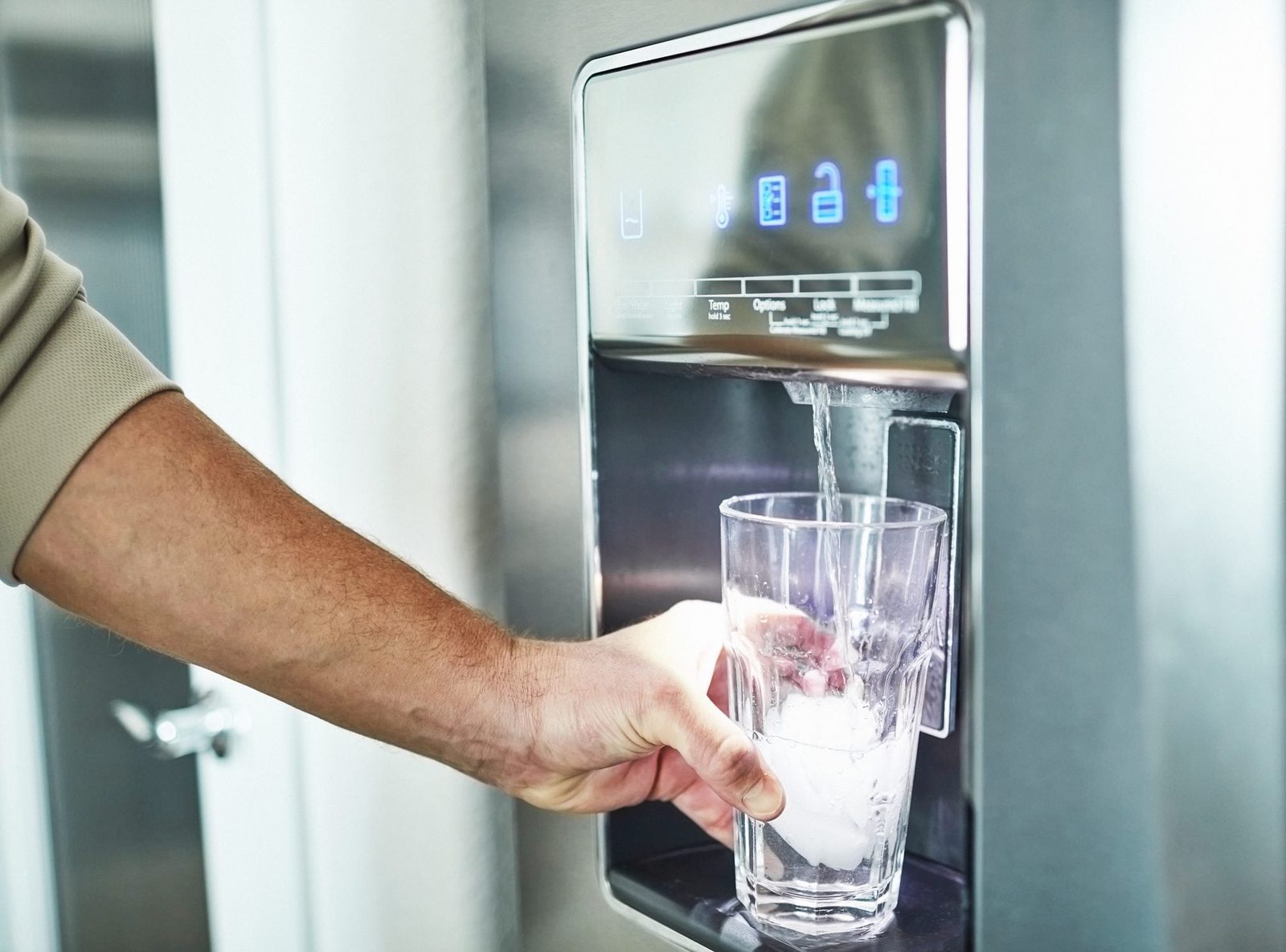 A hand holds a glass under a refrigerator water dispenser, filling it with water over ice in a modern kitchen setting.