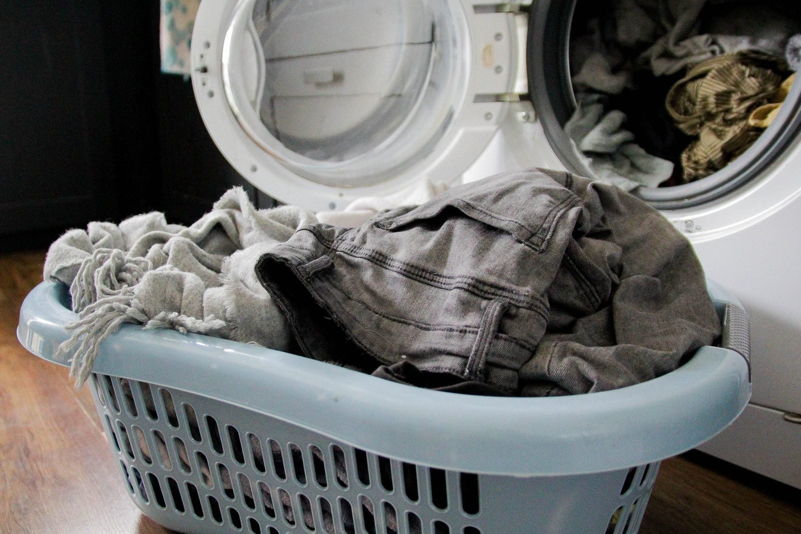 A laundry basket filled with gray clothes sits in front of an open washing machine, surrounded by a wooden floor and dimly lit surroundings.
