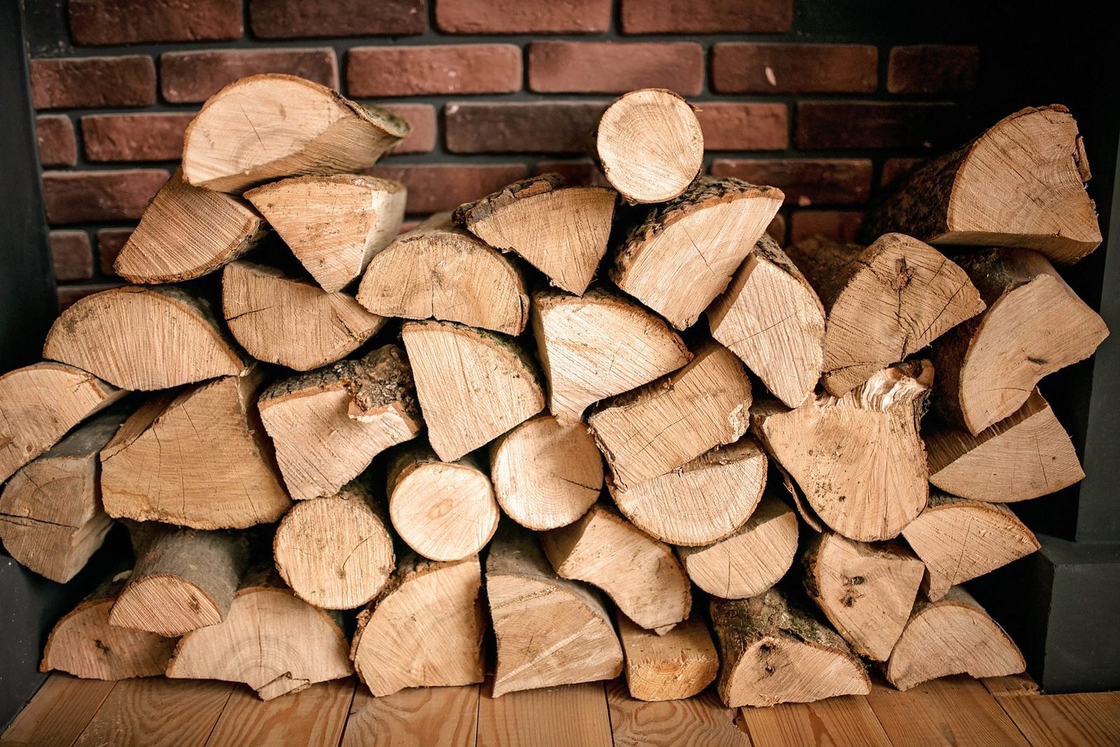 Stacked firewood rests in a cozy setting, showcasing the round ends of logs against a textured brick wall and wooden floor.