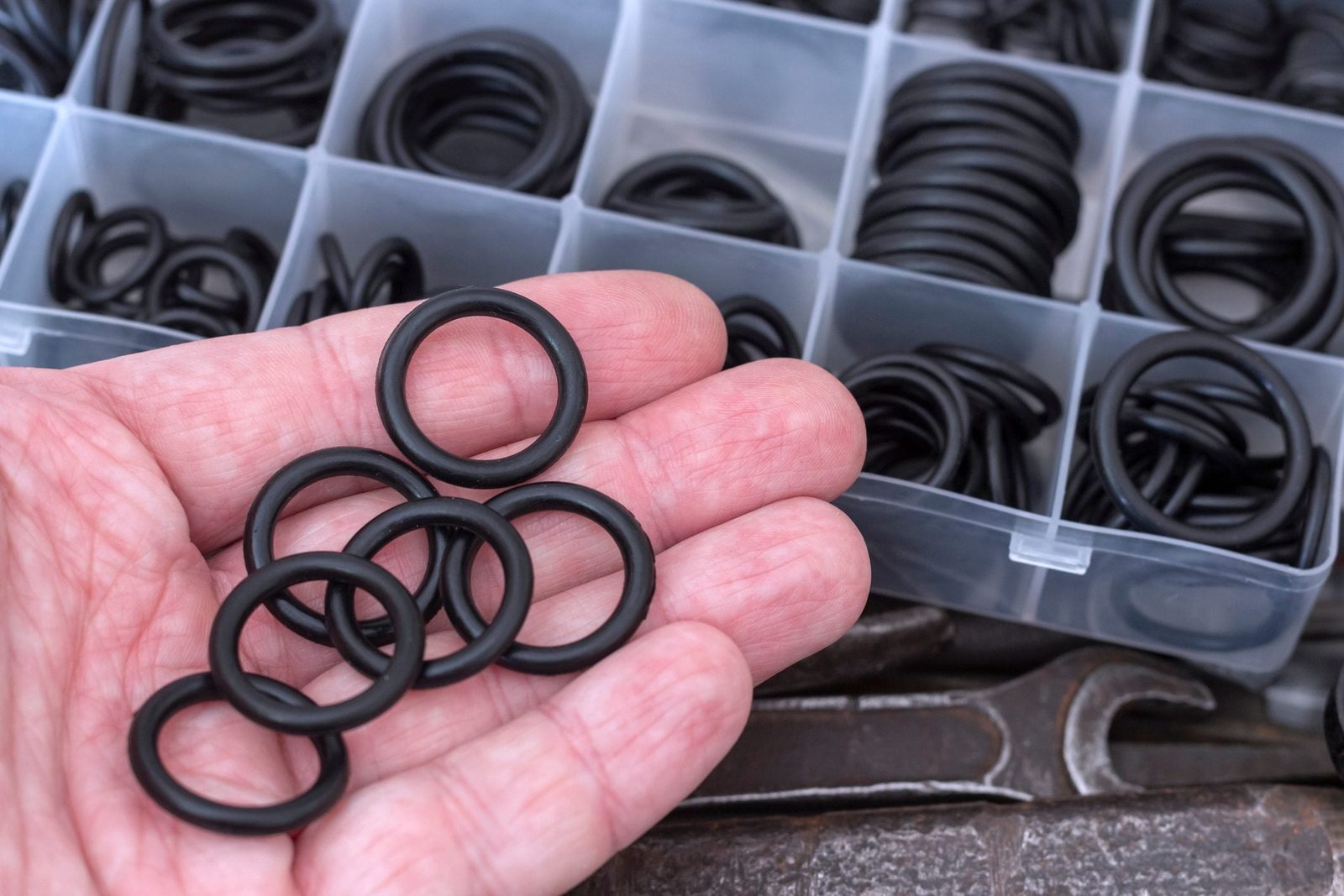 A hand holds several black rubber O-rings while a box of similar rings is visible in the background, resting on a metal surface.