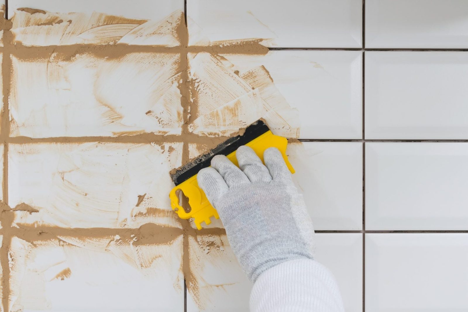 A gloved hand uses a yellow scraper to smooth grout between white tiles on a wall, with uneven, sandy grout visible in the gaps.