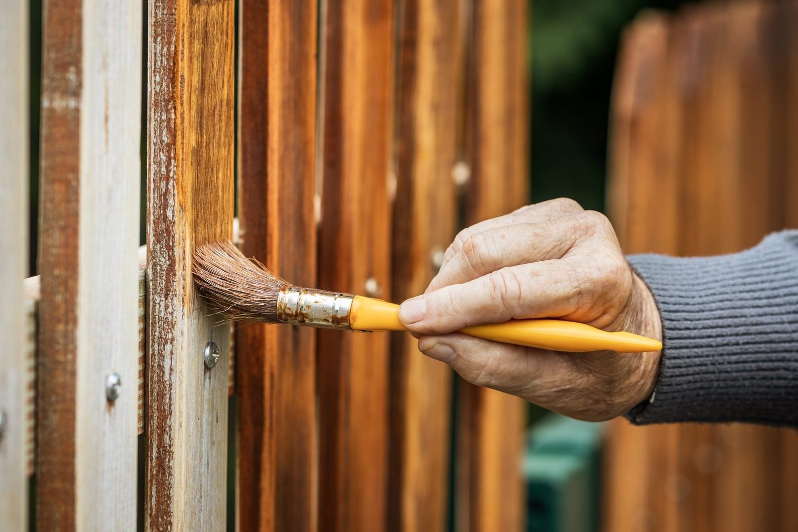 A hand applies a brush with light brown stain to a wooden fence in an outdoor setting, surrounded by vertical wooden panels and lush greener