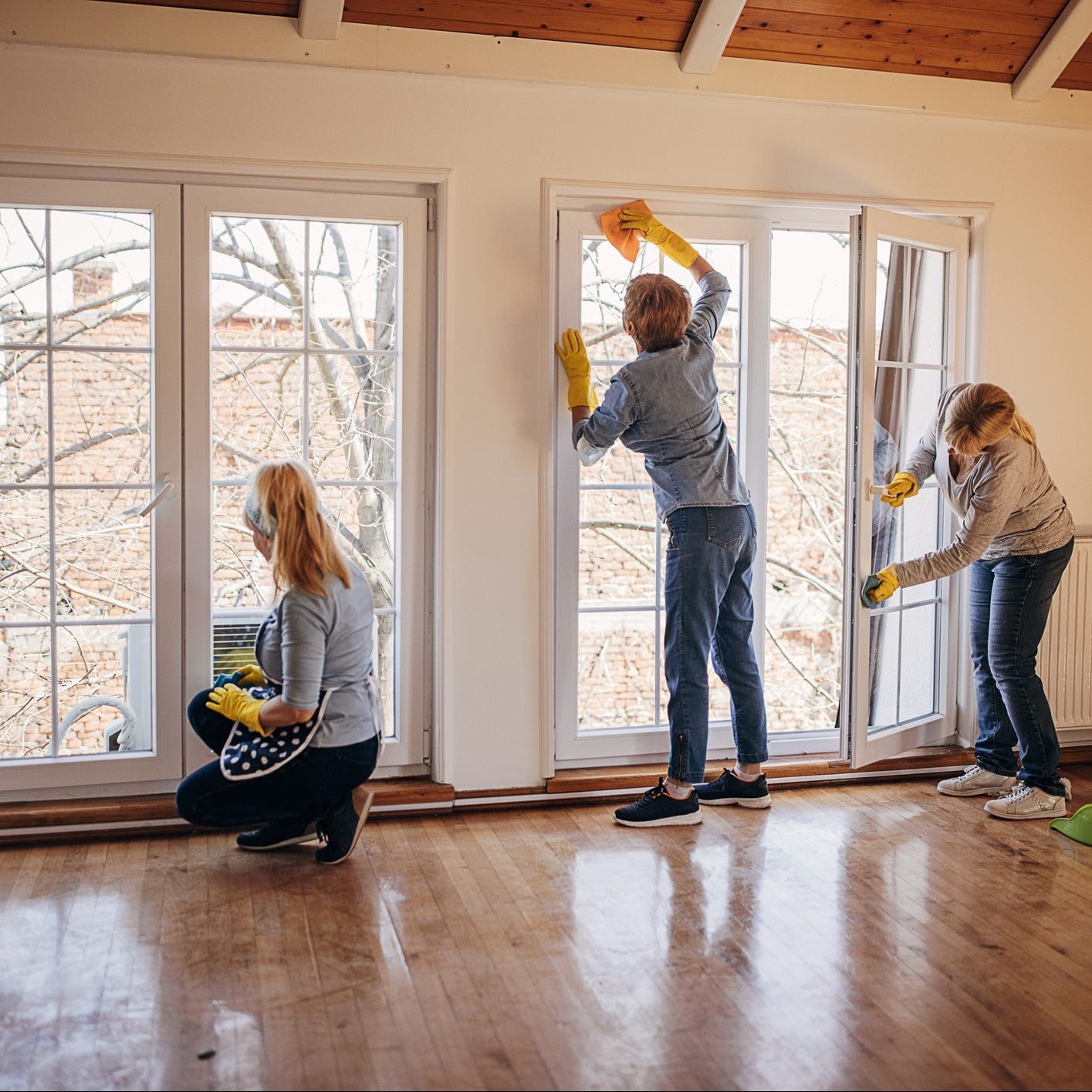 Three people are cleaning windows in a bright, empty room with wooden flooring, wearing gloves and using cloths to wipe the glass surfaces.