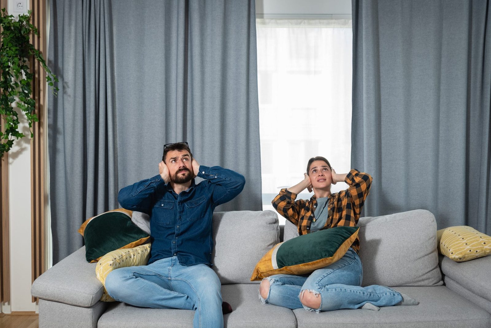 Two people sit on a couch, covering their ears, looking distressed. They are in a bright room with gray curtains and decorative pillows.