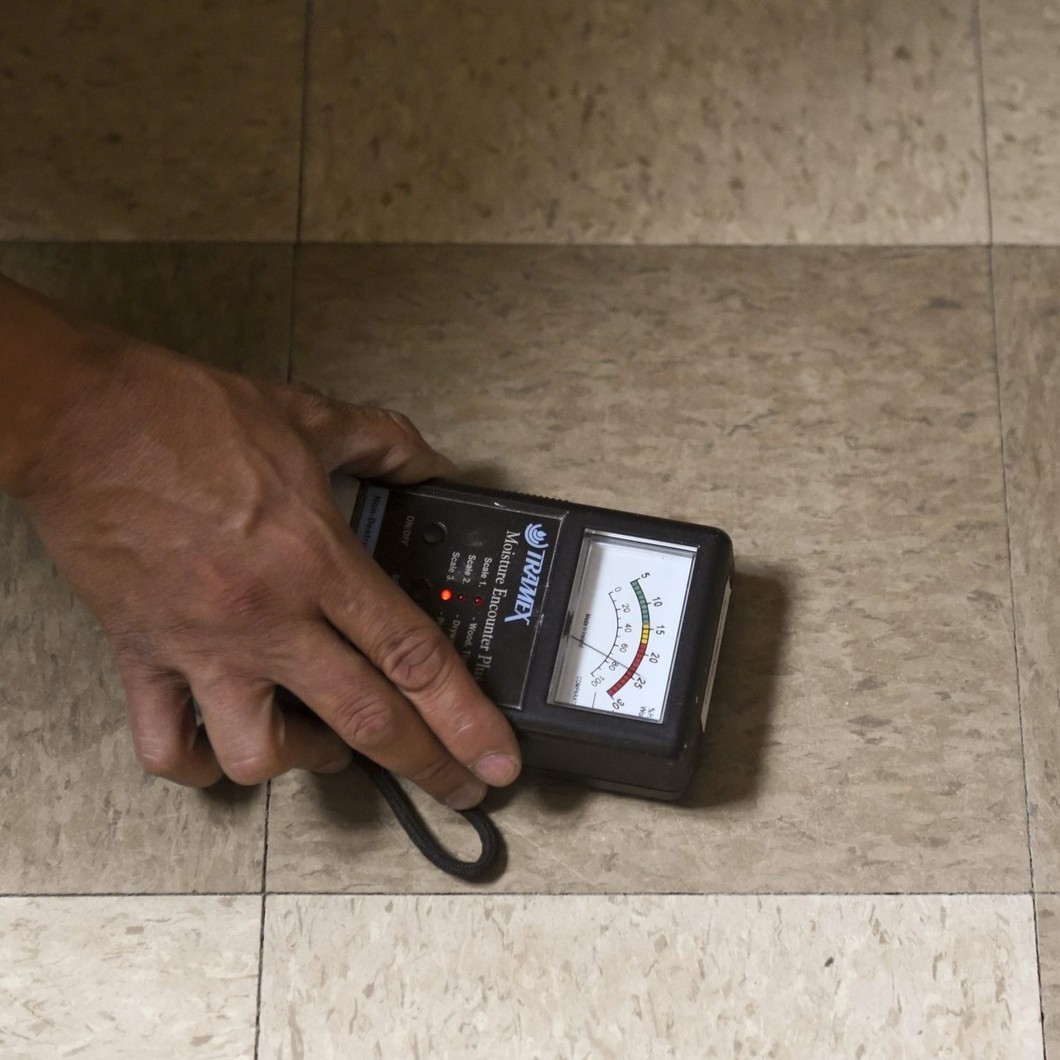A hand holds a moisture meter against a tiled floor, measuring moisture levels in the surrounding environment. The floor has a light-colored