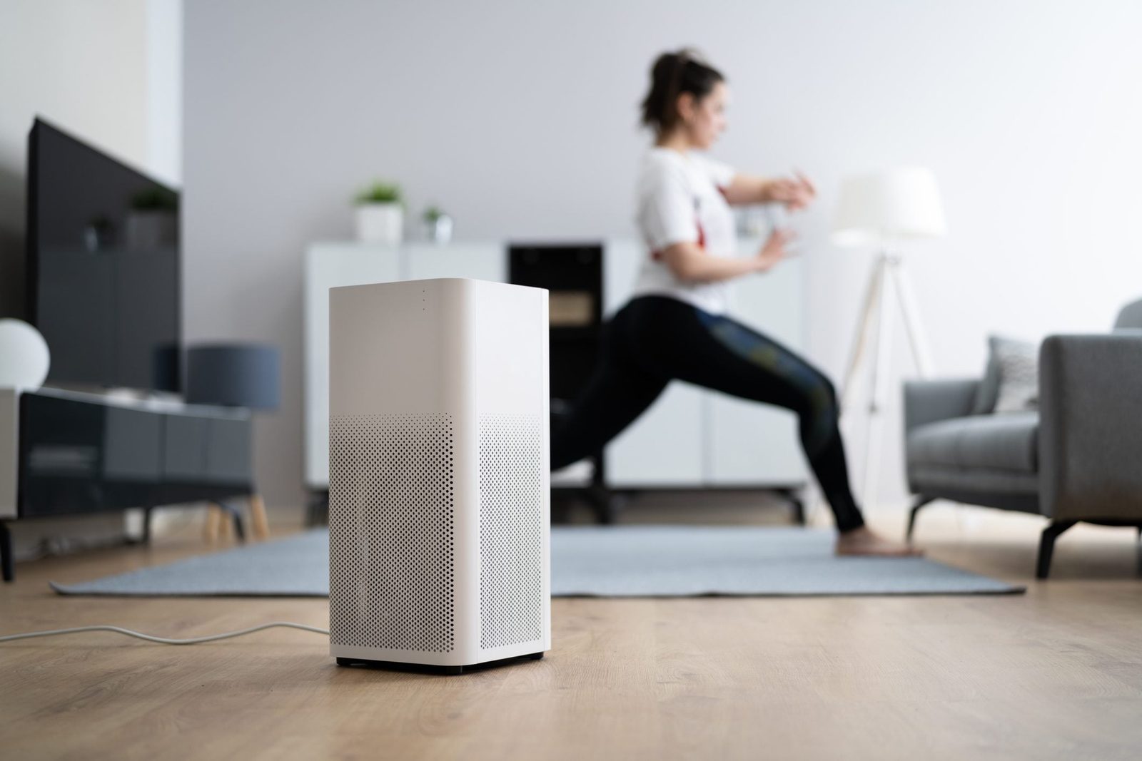 A white air purifier stands on wooden flooring while a person exercises in the background of a modern, well-lit living space.