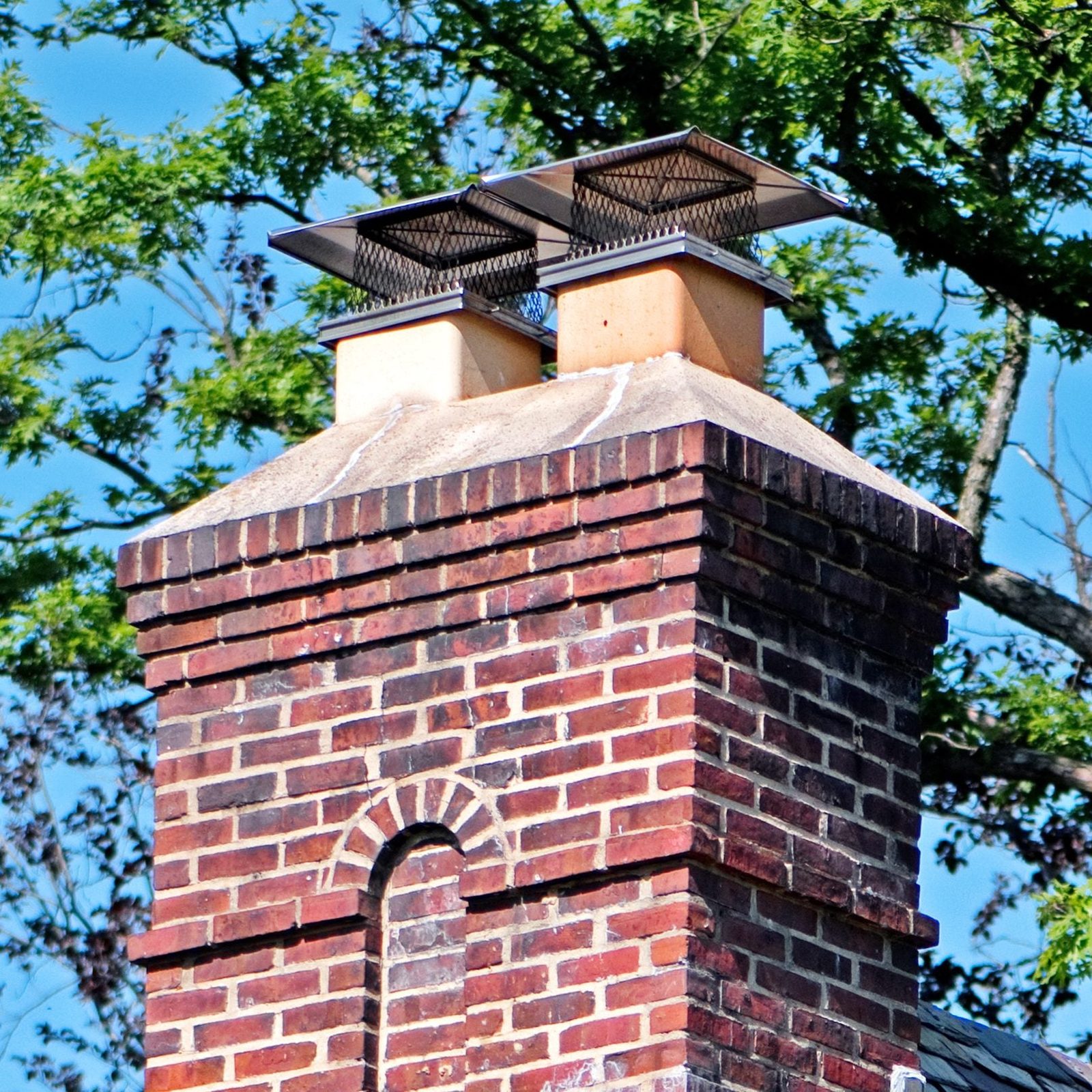 A brick chimney stands tall against a clear blue sky, featuring metal caps on top to prevent debris entry, surrounded by green foliage.