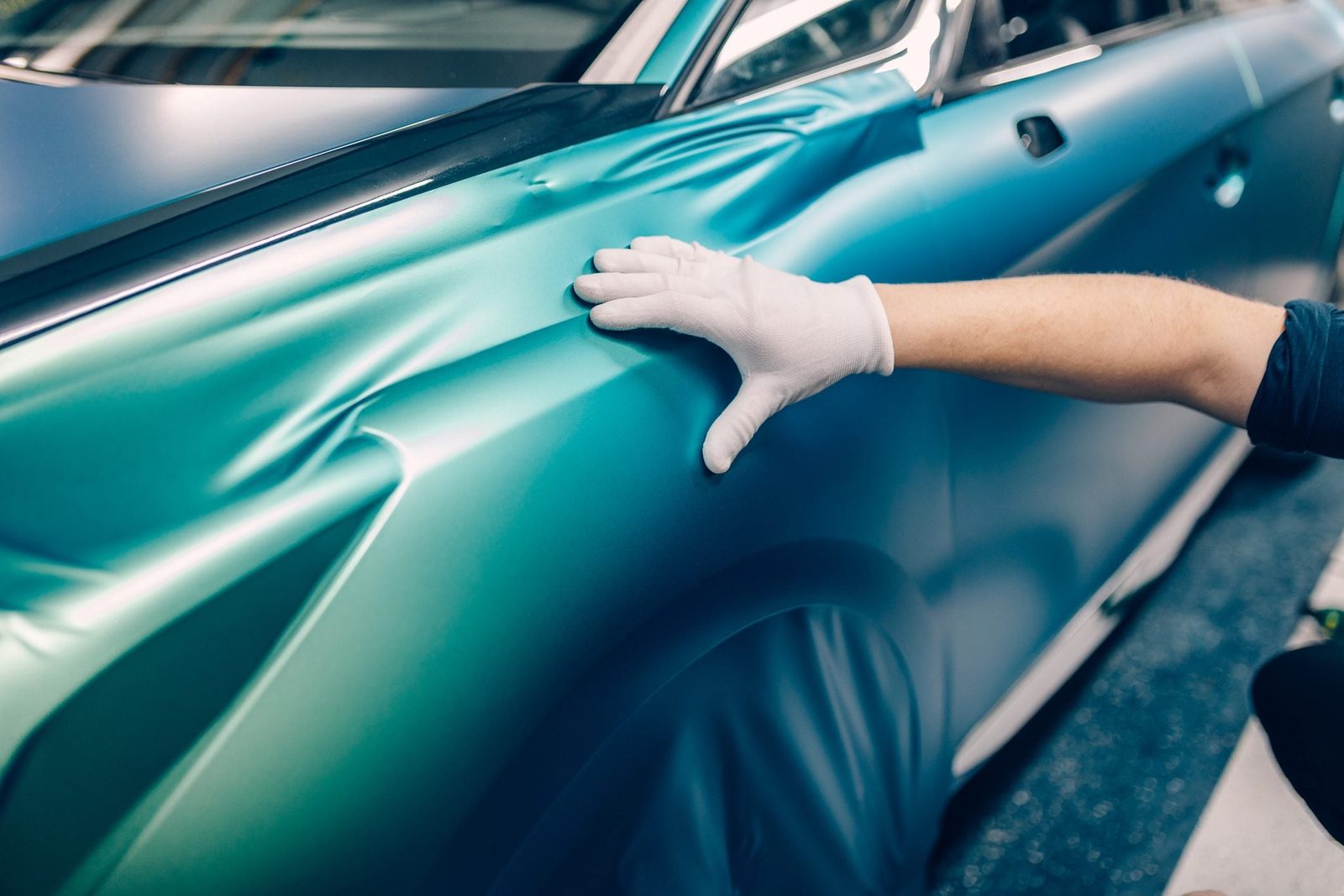 A gloved hand smooths a teal vinyl wrap on a car's side. The setting appears to be an automotive workshop, promoting a clean environment for
