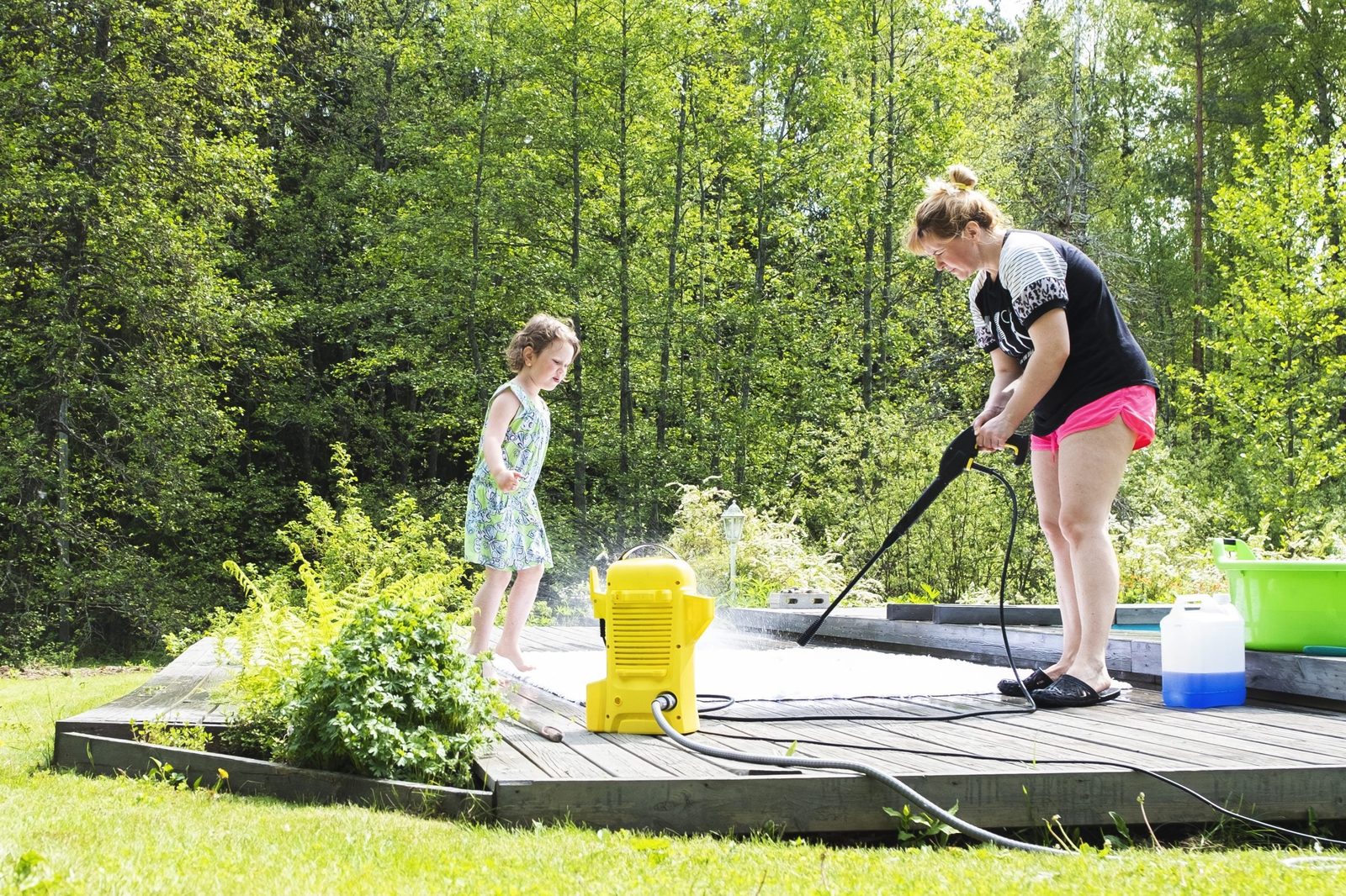 A woman uses a pressure washer while a girl plays nearby on a wooden deck, surrounded by greenery and sunny outdoor scenery.