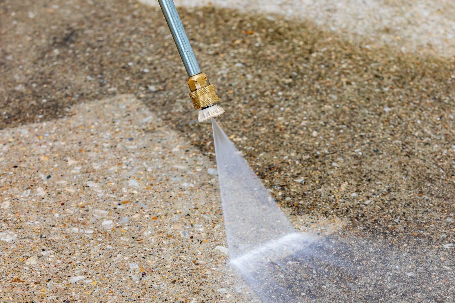A pressure washer nozzle sprays water onto a concrete surface, effectively cleaning dirt while surrounded by a textured gray background.