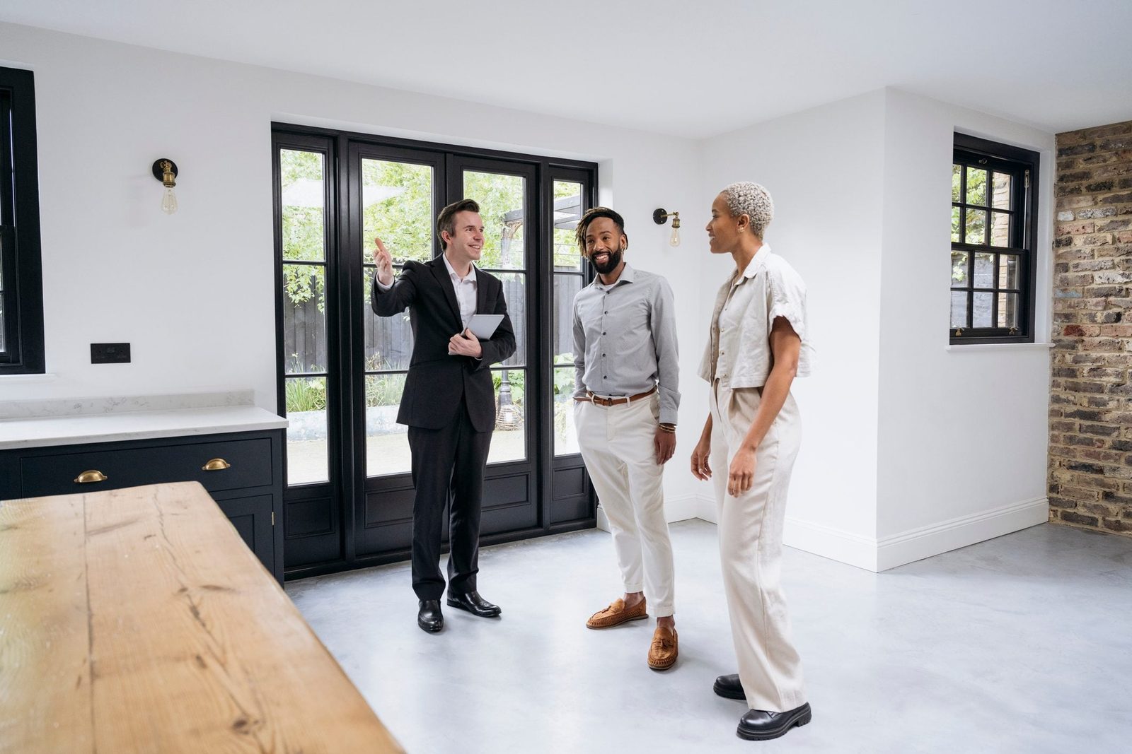 A man in a suit gestures while discussing with two individuals in a modern, well-lit room featuring large windows and a mix of stone and whi