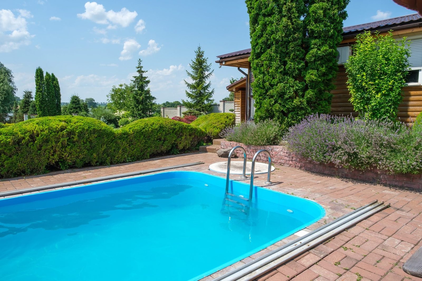 A bright blue swimming pool rests on brick tiles, surrounded by neatly trimmed bushes and vibrant greenery, set against a clear sky and wood