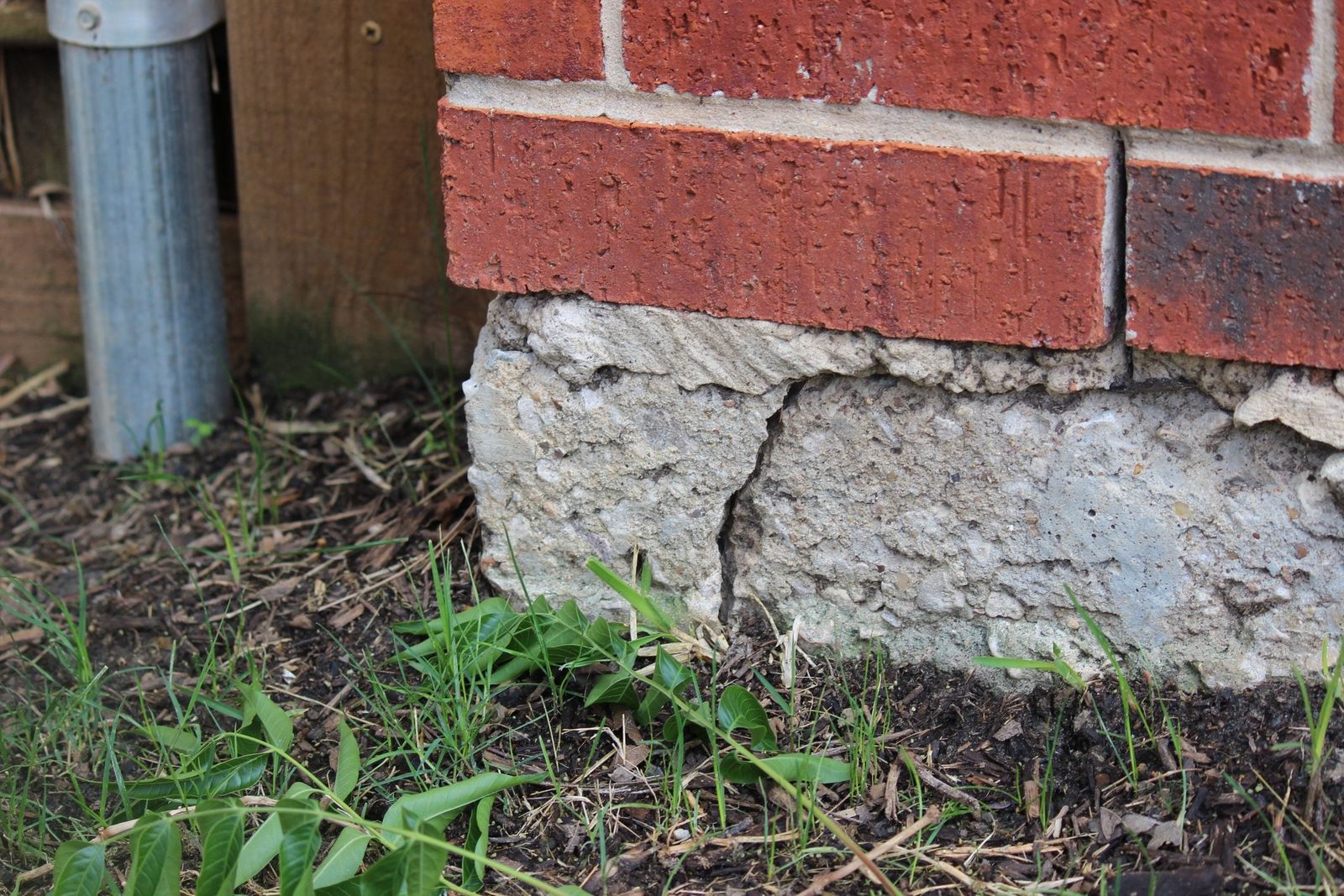 A brick wall stands above a cracked concrete base, surrounded by damp soil and patches of green grass, with a metal pole nearby.