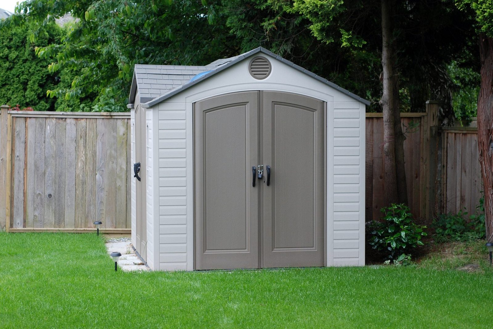 A storage shed stands on green grass, featuring double doors and a gray roof, surrounded by a wooden fence and trees.