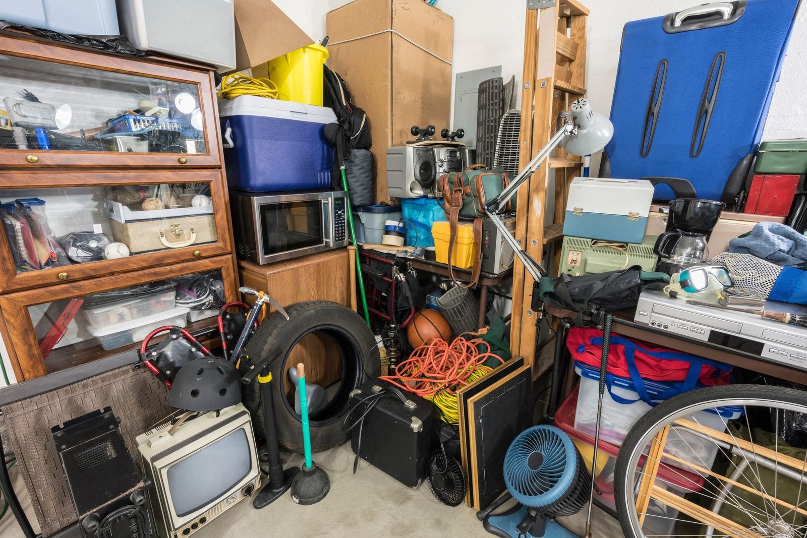 A cluttered garage contains various items like a microwave, bicycle tire, old television, and tools, stacked against walls filled with boxes