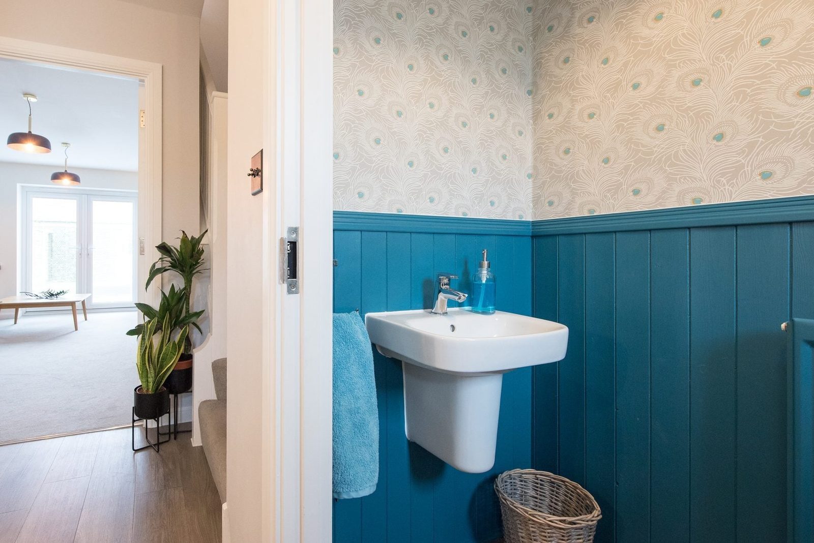 A white sink mounted on a blue-paneled wall, with a decorative bottle and towel, near a doorway leading to a bright, open living space.