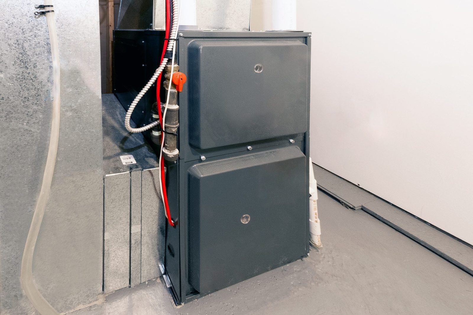 A gray furnace sits against a wall, connected to pipes and wires, in a basement with a concrete floor and minimal lighting.