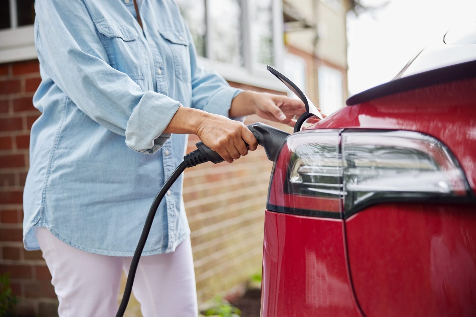 A person connects a charging cable to the rear of a red electric vehicle in a residential setting, with brick walls and windows visible in t