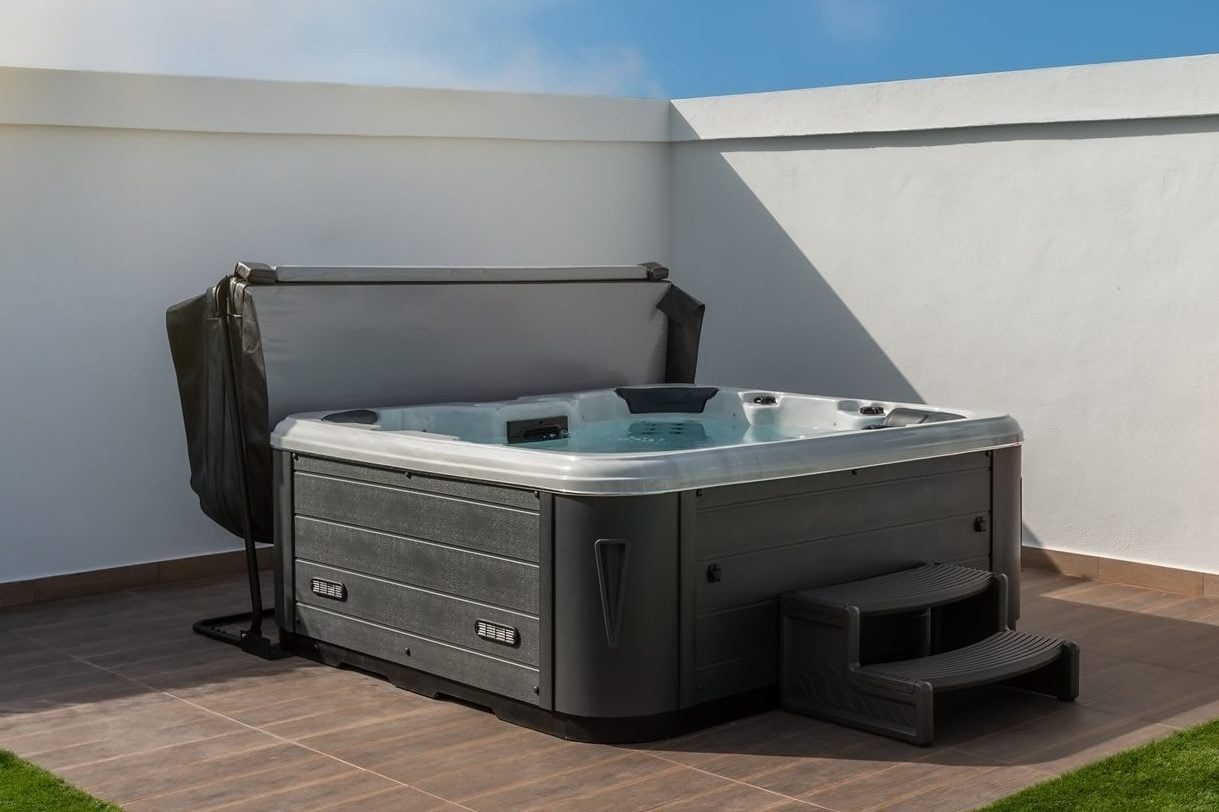 A hot tub sits on tiled flooring, with a cover partially lifted, surrounded by white walls and a patch of green grass under a clear blue sky