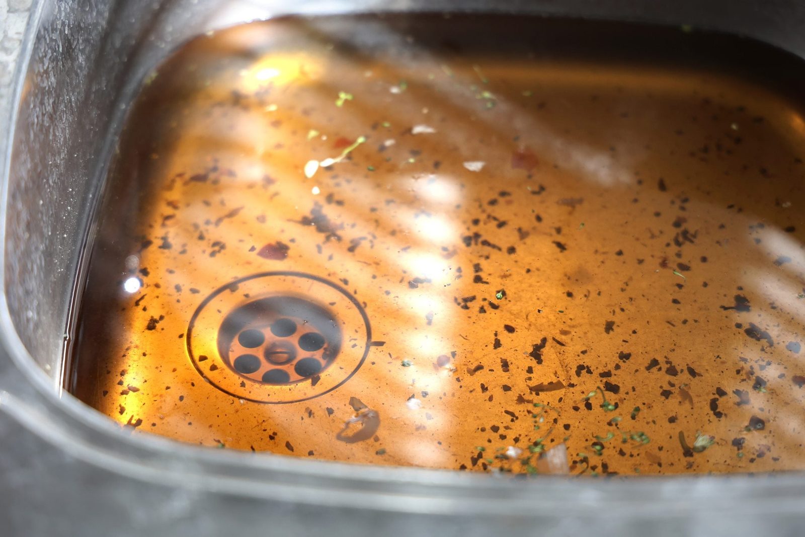 A kitchen sink contains murky, brown water filled with debris and food particles, reflecting light and surrounding a circular drain at the b