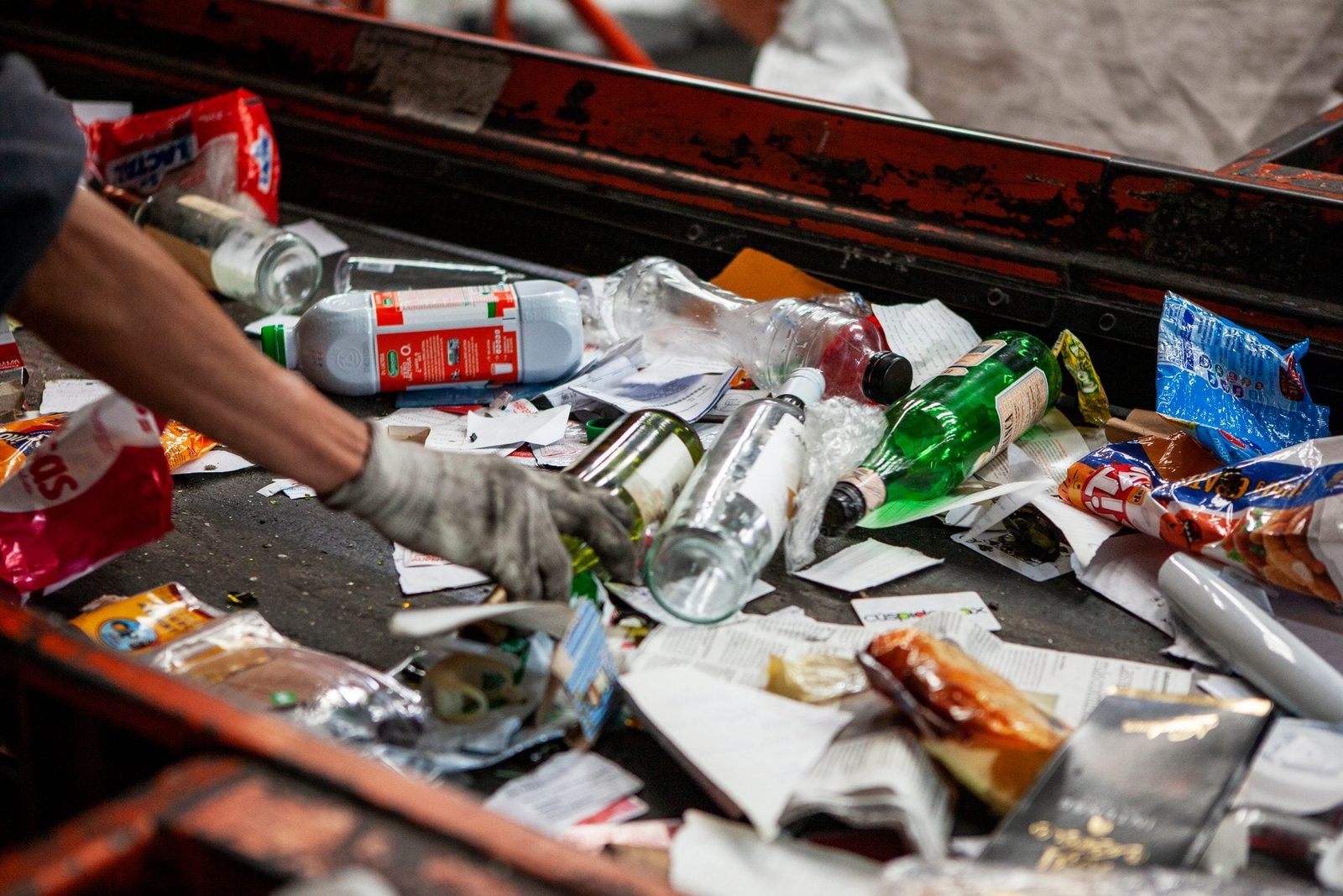 A gloved hand sorts through various discarded items on a conveyor belt, including bottles and food packaging, in a waste processing facility