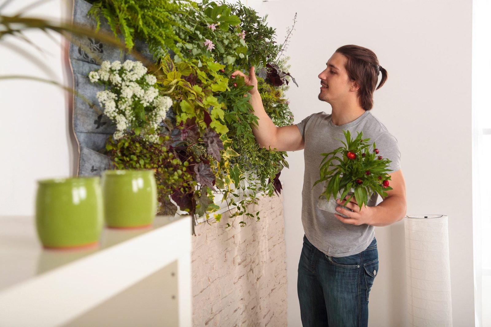 A man arranges plants on a vertical garden while holding a potted plant, surrounded by a bright, modern indoor space.
