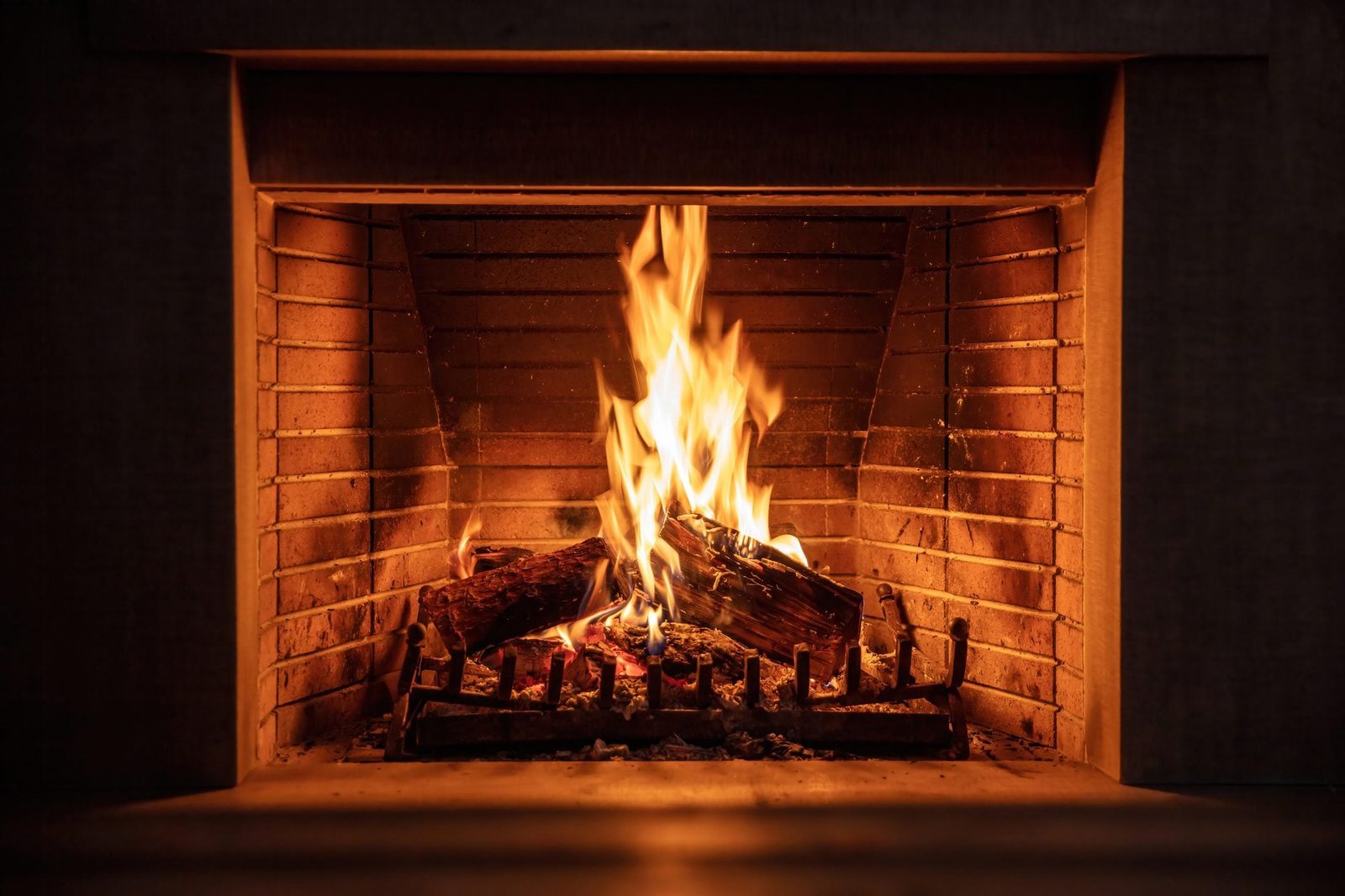 A fire burns brightly in a brick fireplace, casting warm light and flickering shadows in a cozy indoor setting. Logs crackle on the grate.