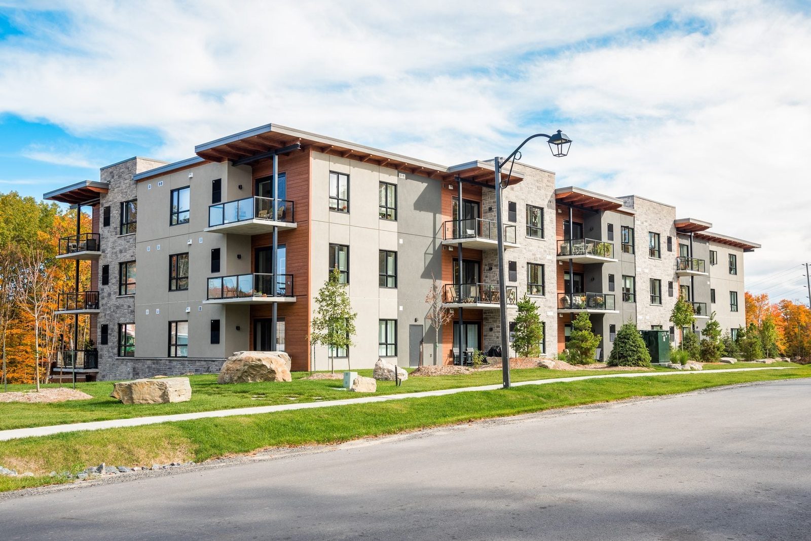 A modern apartment building features multiple balconies, surrounded by grass and trees, with a sidewalk and street in front, under a partly