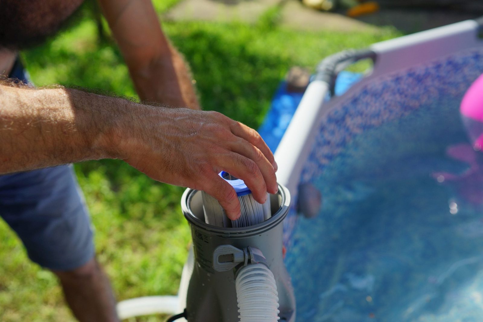 A hand places a filter into a pool cleaner, positioned beside a blue inflatable pool on a sunny day with green grass in the background.