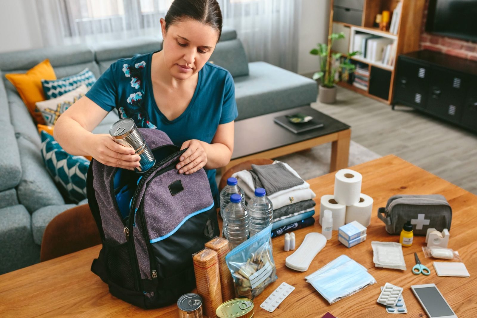 A woman packs a backpack with canned food and various supplies while sitting at a table in a cozy living room.
