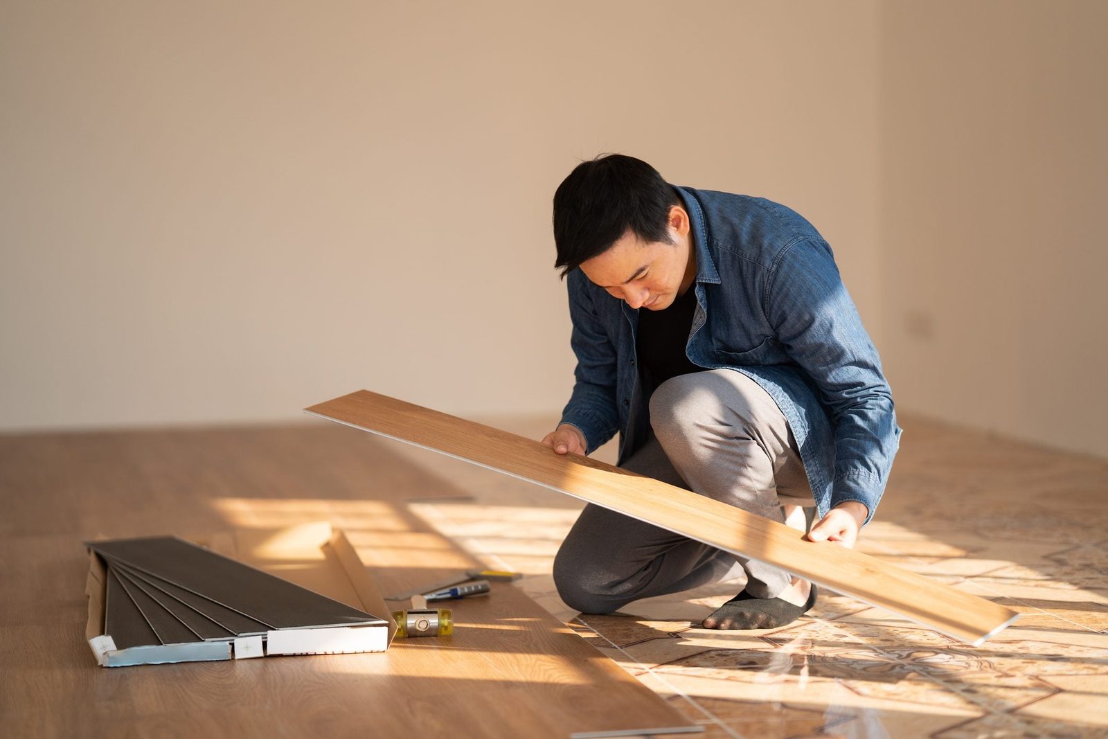A man examines a wooden plank while kneeling on the floor. Tools and additional planks are nearby in a bright, empty room.