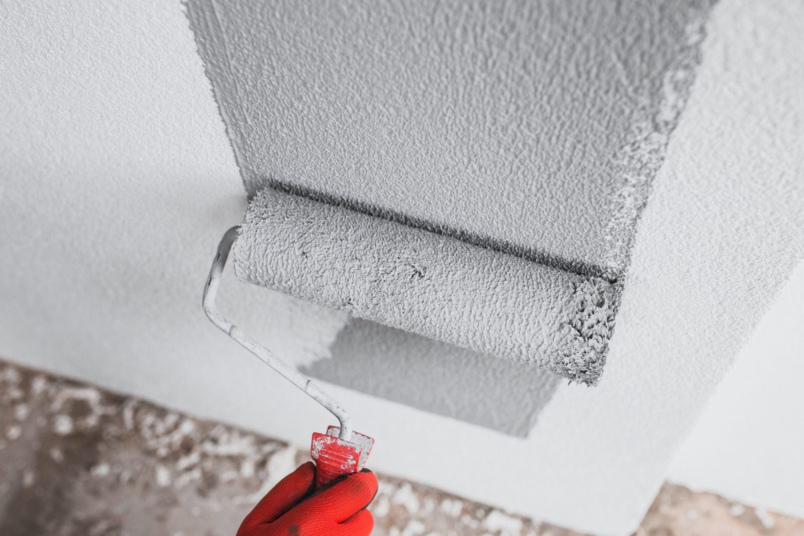 A hand with a red glove holds a paint roller, applying gray texture paint to a wall in a partially finished room.