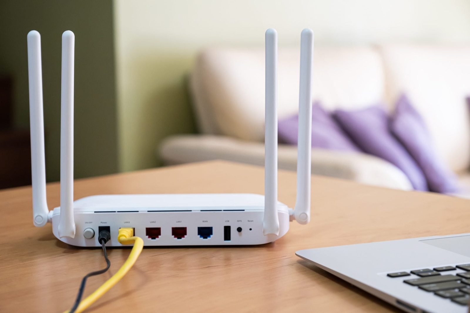 A white router with two antennas sits on a wooden table, connected to cables and a laptop nearby, in a cozy indoor environment.