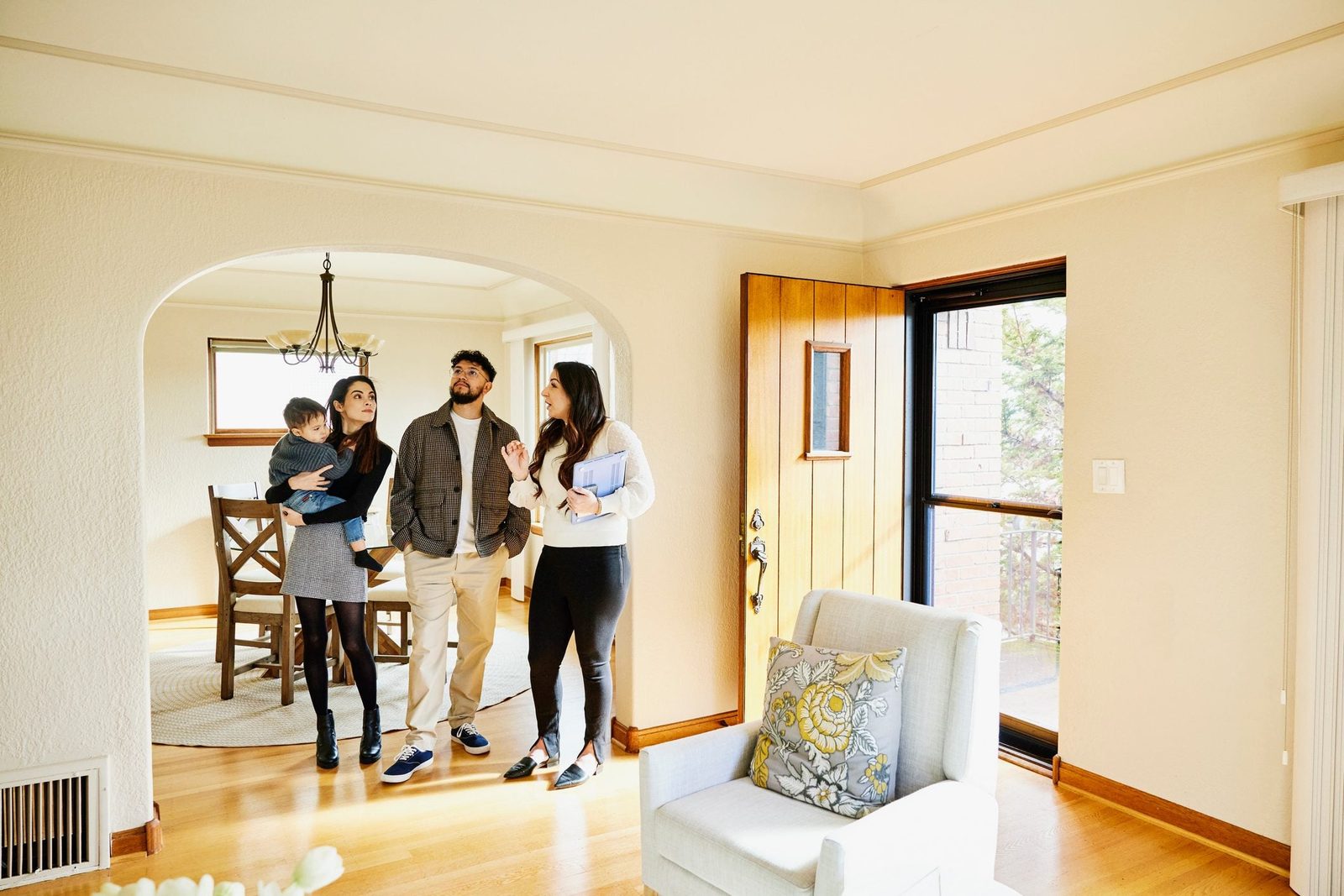 A family tours a bright living room, discussing with a guide while admiring the space. Wooden furniture and a large window add warmth to the