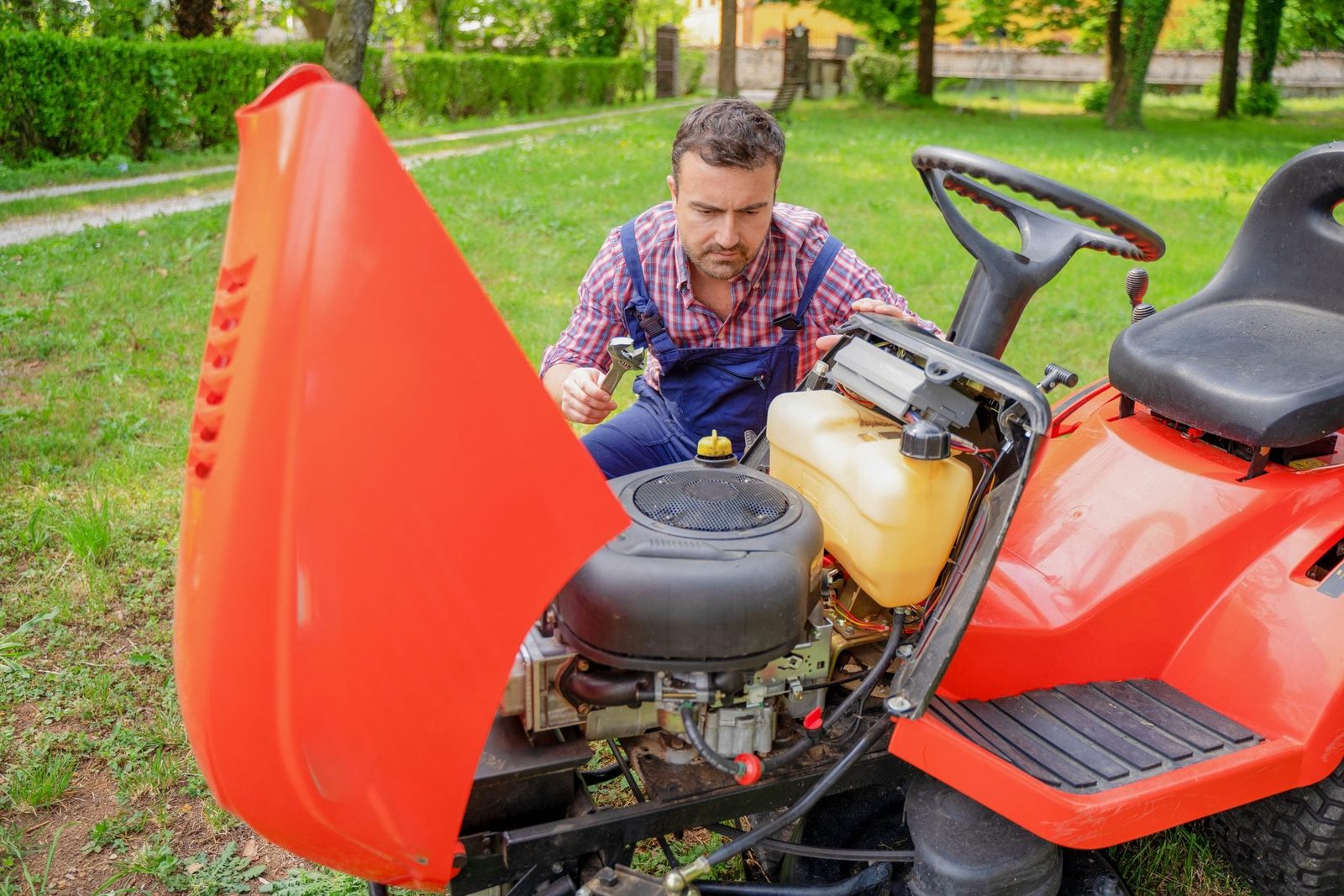 A person inspects a small engine under the hood of a red riding lawn mower, surrounded by green grass and trees in a backyard.