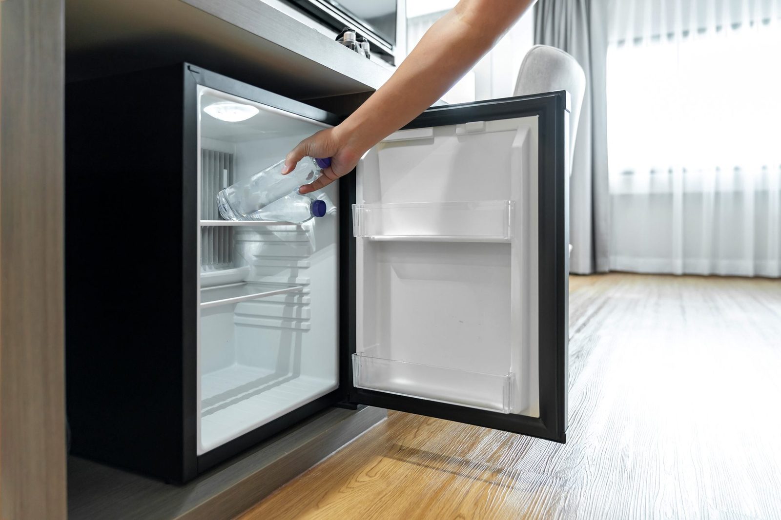 A hand is placing a water bottle into a small refrigerator, situated next to a wooden surface in a well-lit room with curtains.