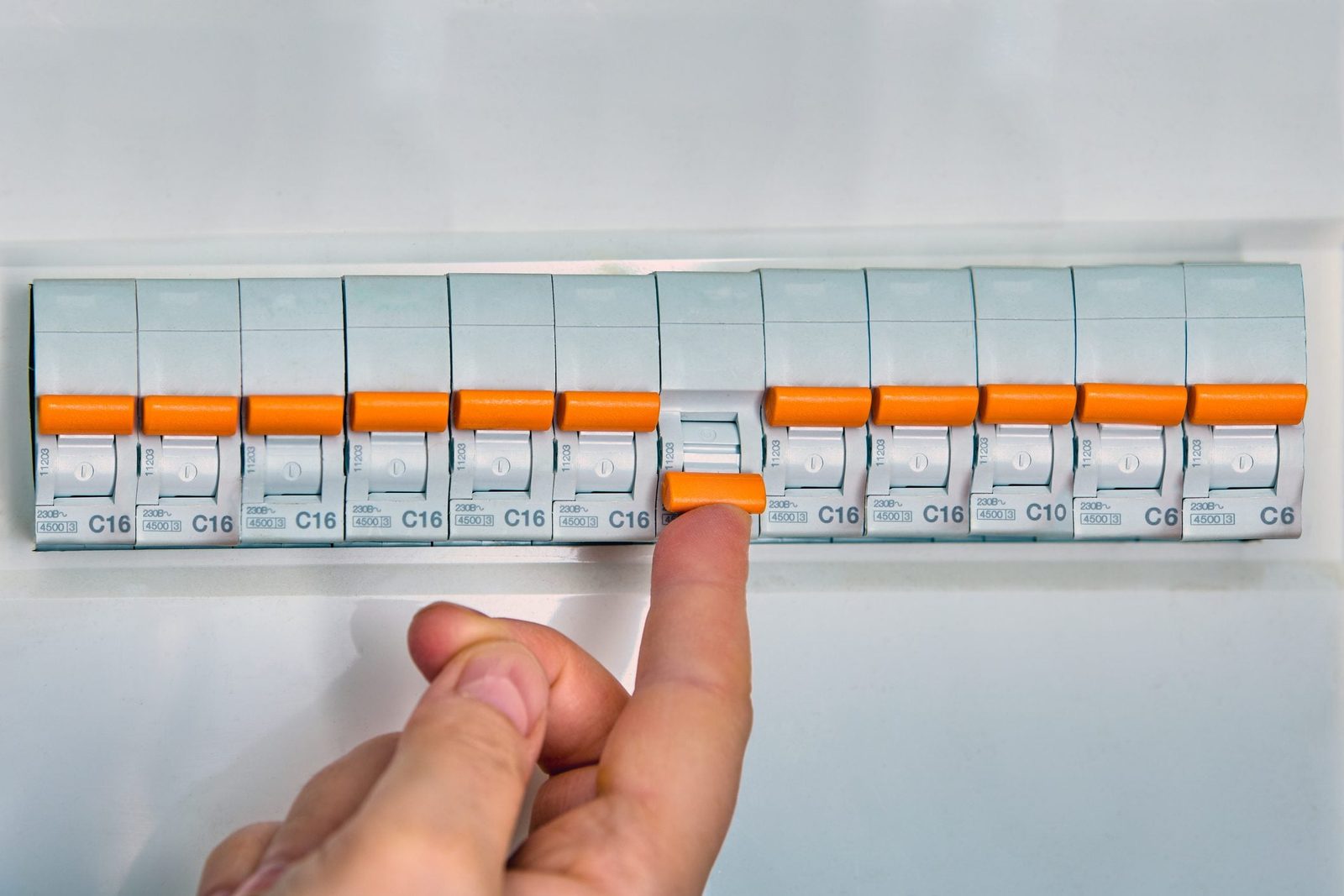 A hand is pressing down a switch on a circuit breaker panel, surrounded by multiple switches labeled with numbers and letters, indicating el