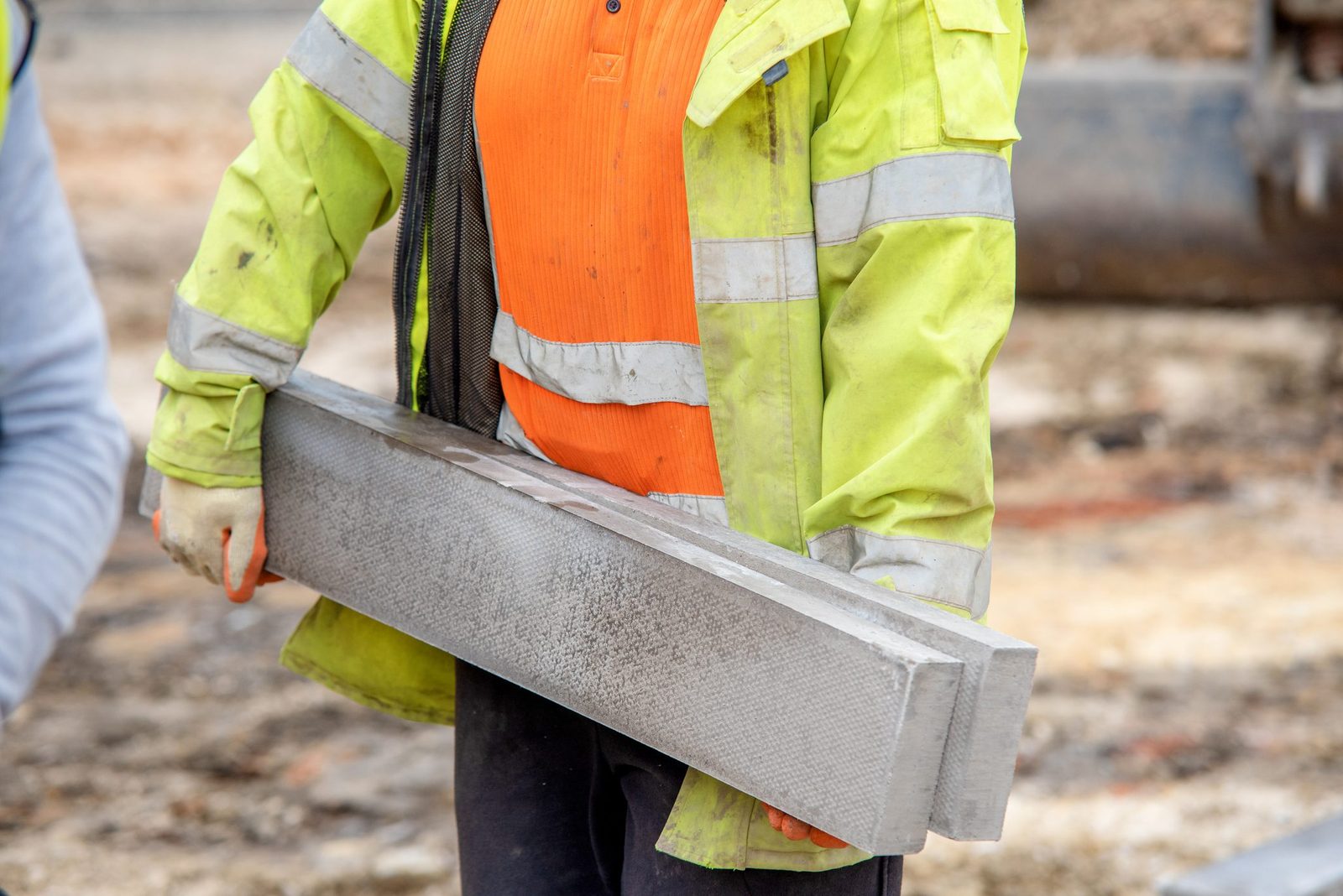 A construction worker holds a large concrete block, wearing a bright yellow jacket and orange shirt, amid a rough, unfinished outdoor worksp