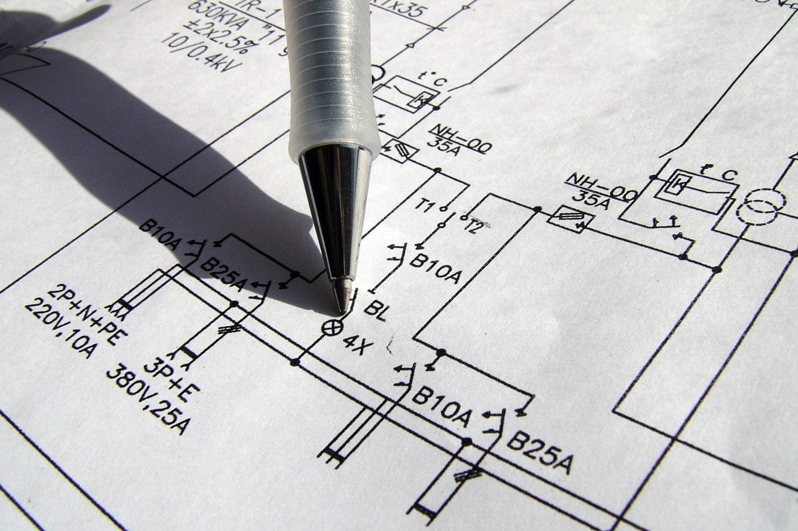 A silver pen points at a detailed electrical schematic on white paper, showcasing various electrical symbols and connections under natural l