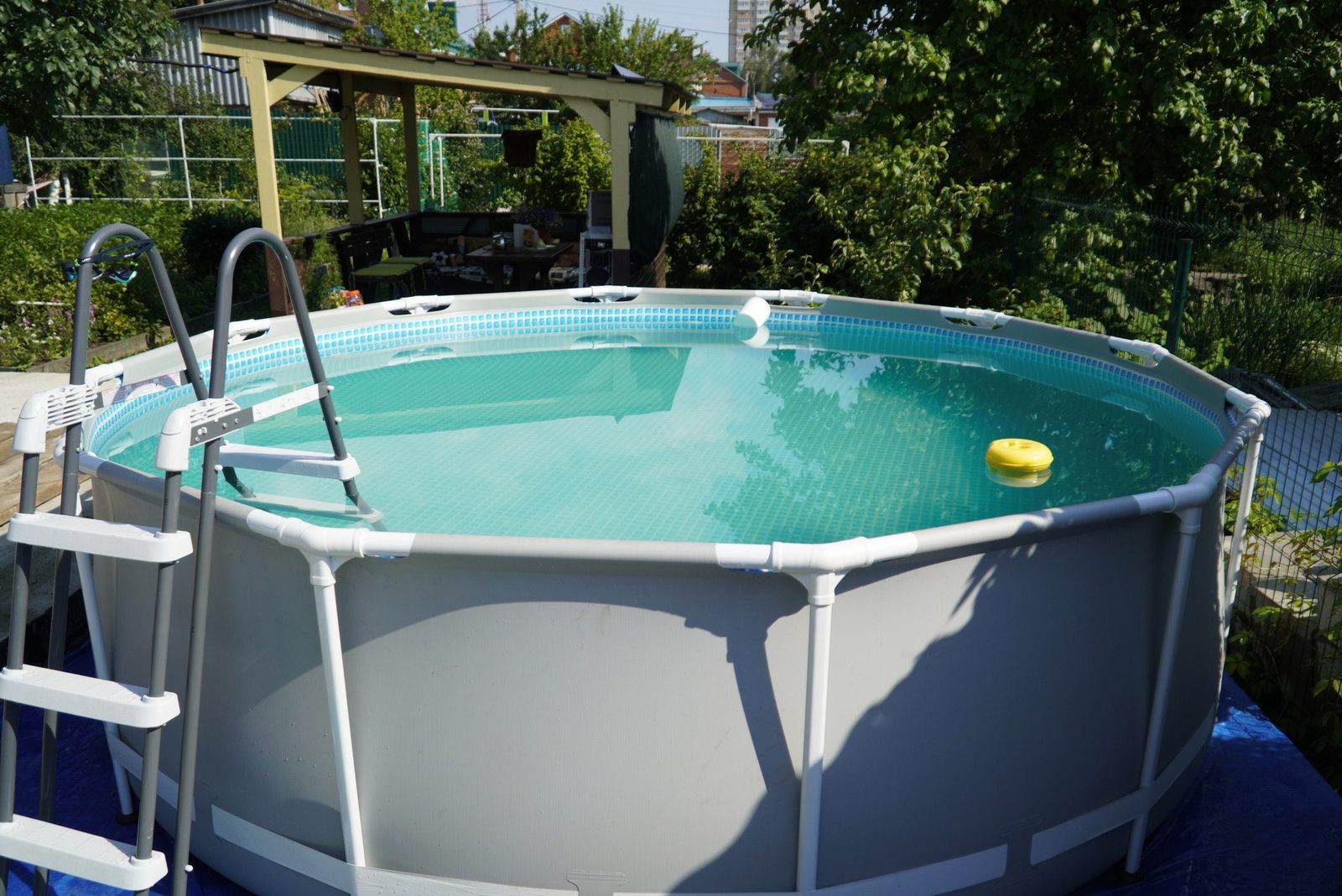 A round above-ground swimming pool holds clear water, with a yellow float visible. A ladder leans against the pool in a garden setting.