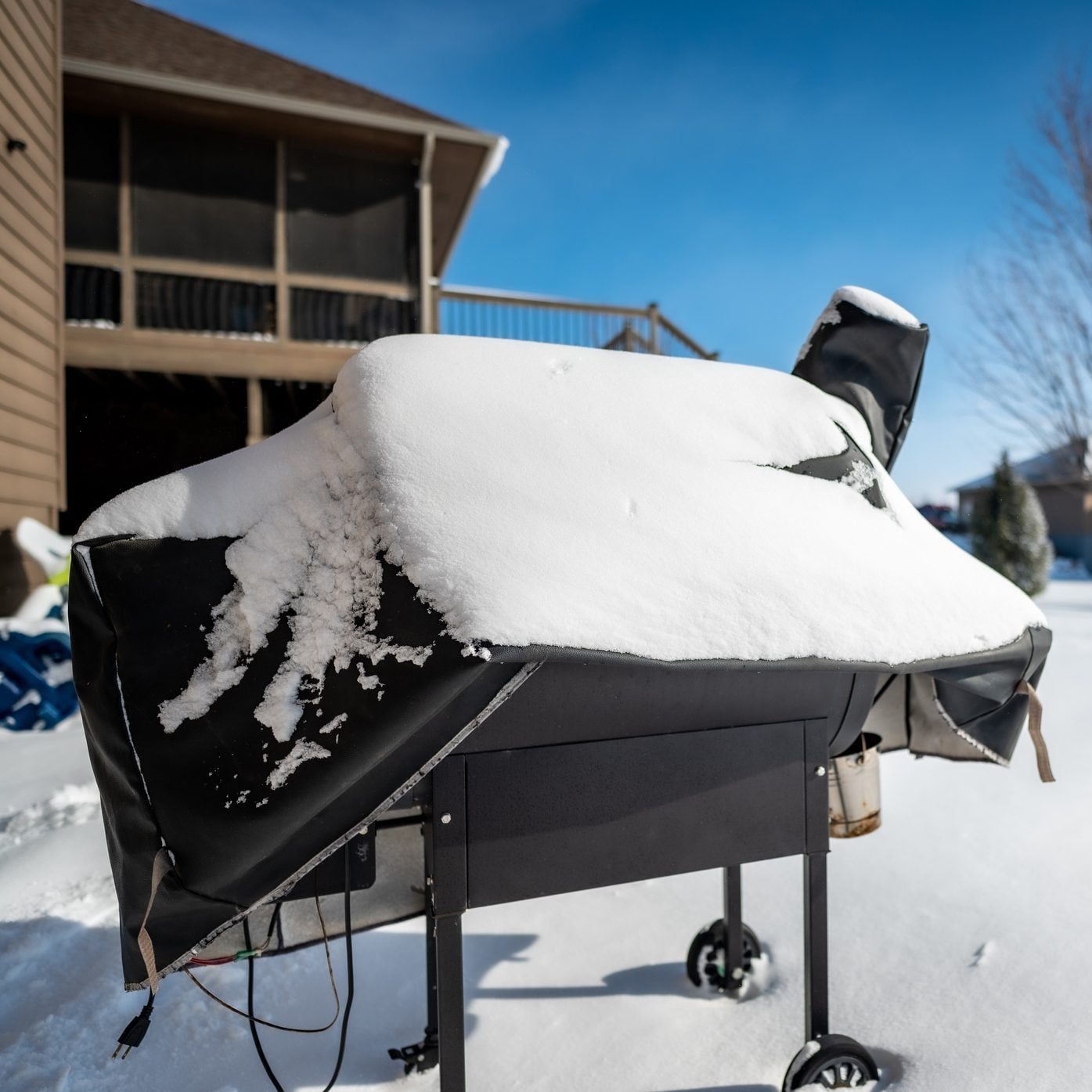 A grill covered in snow sits on a snowy yard, with a clear blue sky above and a house with a screened porch in the background.