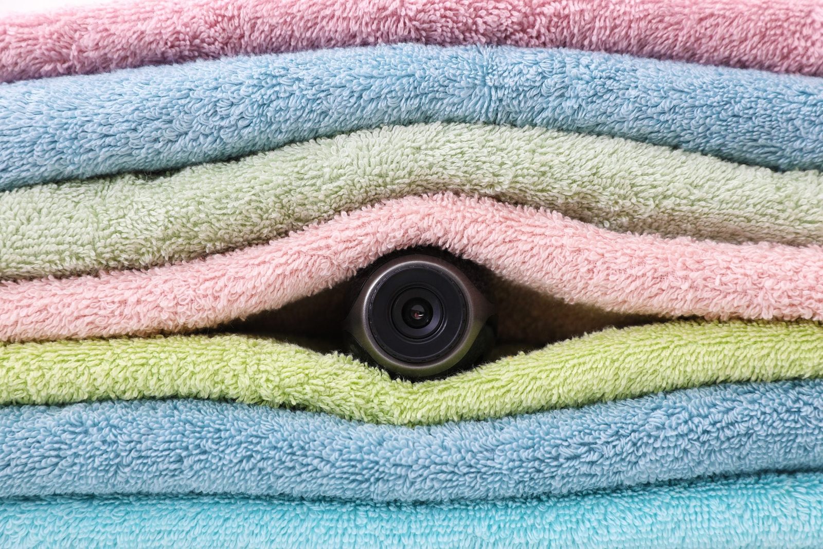 A camera lens peeks out from between stacked, colorful towels, showcasing a cozy, neatly organized bathroom or laundry environment.