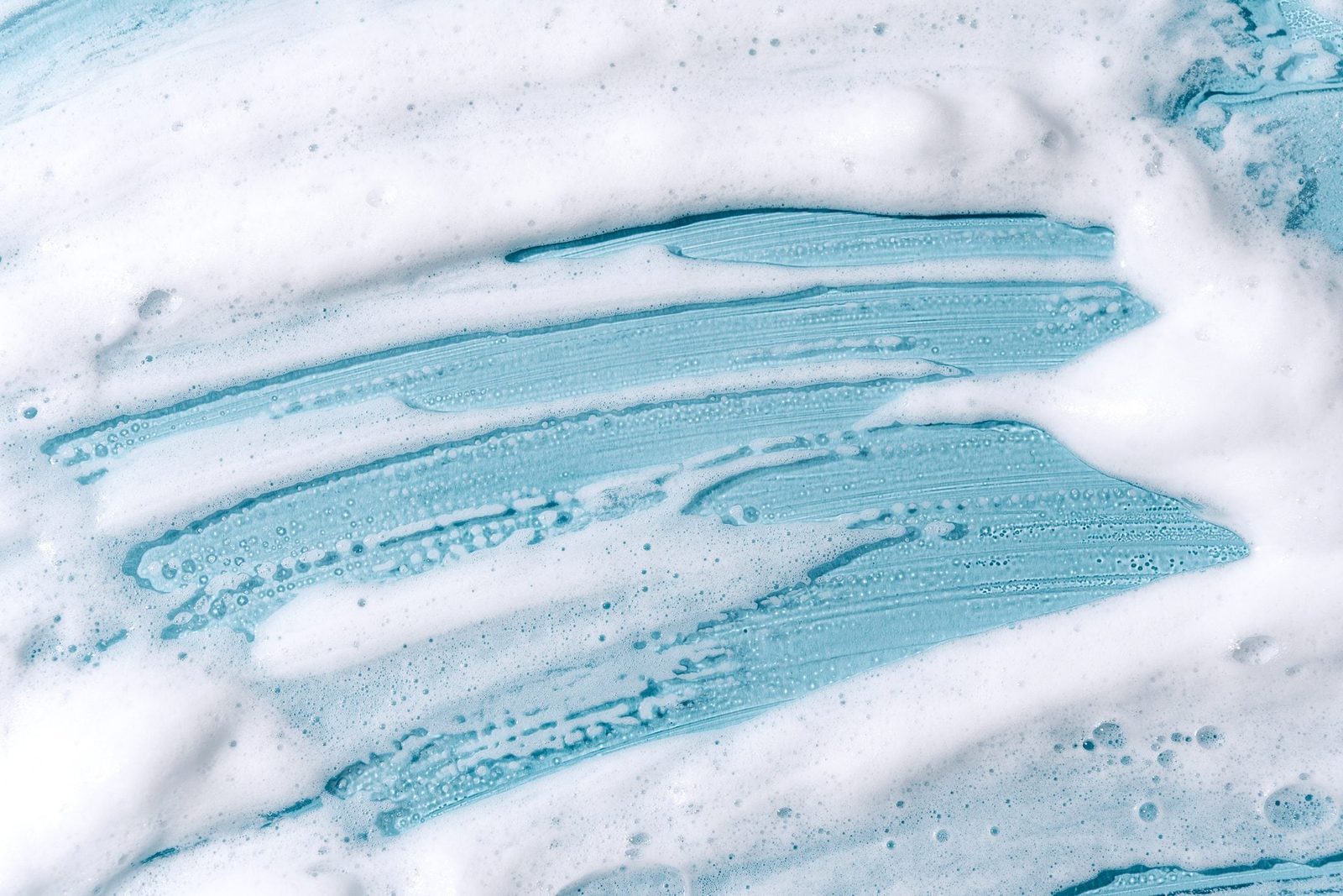 A blue cleaning solution creates streaks and bubbles while being spread across a white surface, suggesting a cleaning context.