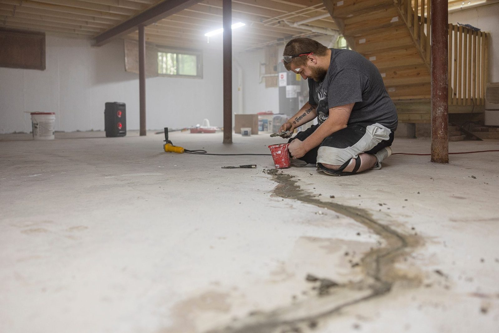 A man kneels on a concrete floor, applying a substance from a bucket, in a spacious, unfinished basement with visible construction materials