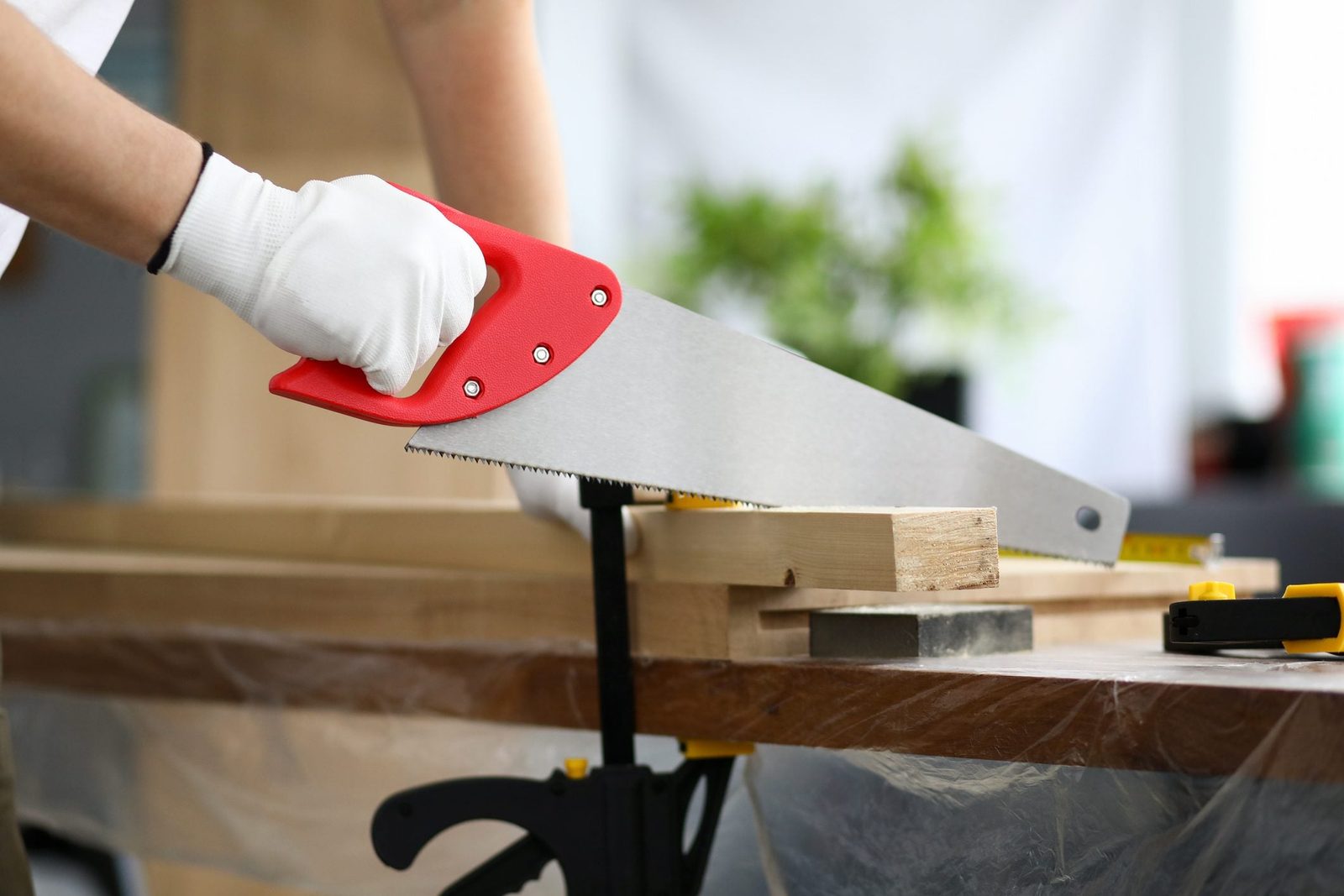 A gloved hand holds a red-handled saw, cutting through a wooden plank secured on a workbench, surrounded by a blurred indoor environment.